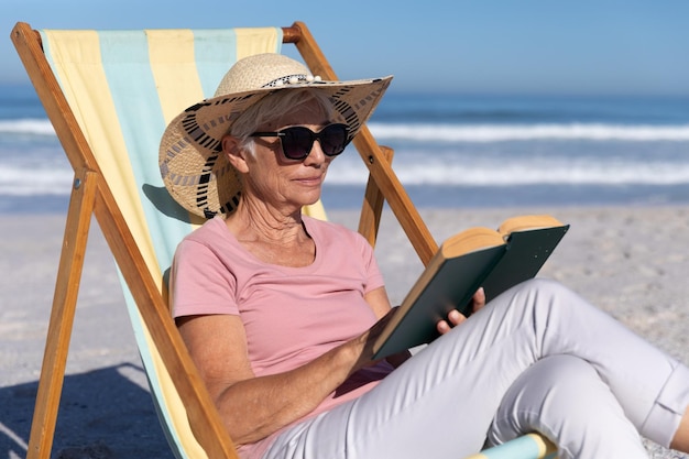 Mulher caucasiana sênior aproveitando o tempo na praia, sentada em uma espreguiçadeira e lendo um livro