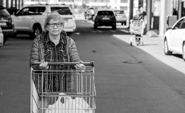 Foto mulher caucasiana sênior com carrinho de compras vai para o estacionamento do supermercado foto em preto e branco