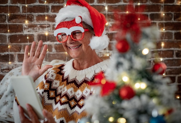 Mulher caucasiana sênior feliz com óculos engraçados e suéter de inverno usando vídeo por telefone conversando com família ou amigos senhora idosa comemora o Natal e o ano novo em casa