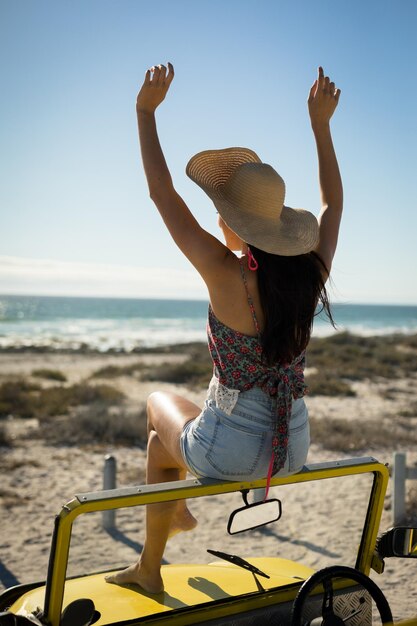 Foto mulher caucasiana sentada em um buggy de praia ao lado do mar usando um chapéu de palha olhando para o mar com as mãos levantadas