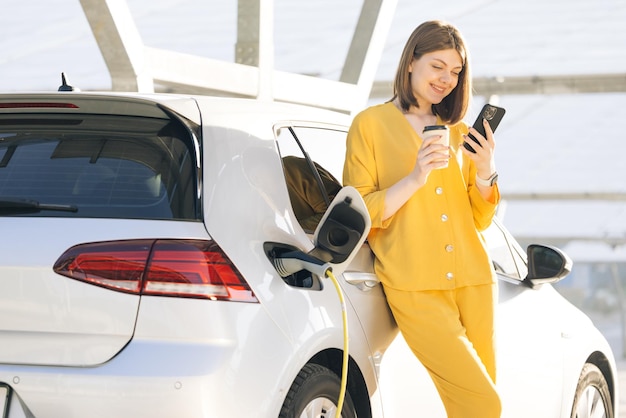 Mulher caucasiana usando telefone inteligente e fonte de alimentação em espera conecta-se a veículos elétricos para carregamento