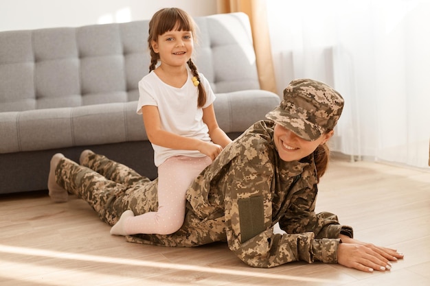 Mulher caucasiana, vestindo uniforme de camuflagem e boné brincando com sua filha depois de voltar para casa do exército, linda filha sentada nas costas da mãe enquanto a mãe deitada no chão.