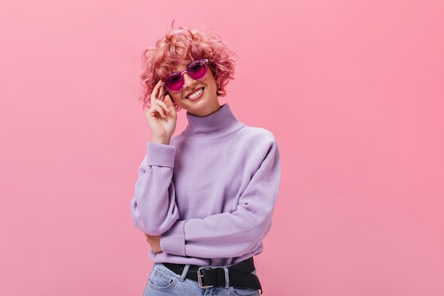 Mulher charmosa de cabelo curto com óculos escuros fúcsia e suéter roxo com um sorriso largo na parede rosa