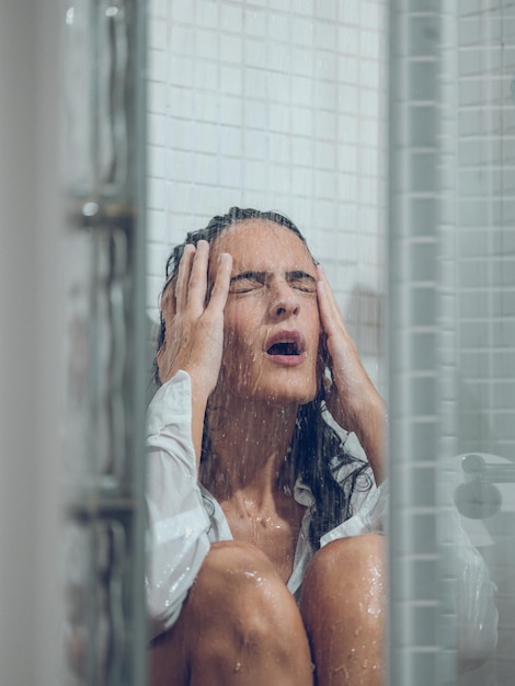 Mulher chateada com cabelos escuros e camisa branca molhada chorando enquanto está sentada no chuveiro sob o riacho com os olhos fechados e tocando a cabeça