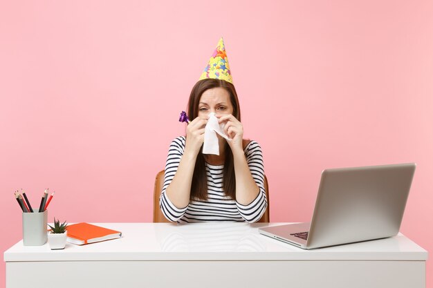 Mulher chateada com chapéu de festa com cachimbo enxugando as lágrimas com lenço, porque ninguém veio comemorar na mesa branca com laptop pc isolado no fundo rosa. Carreira empresarial de realização. Copie o espaço.