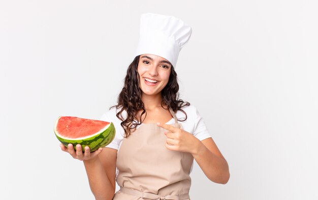 mulher chef bonita hispânica parecendo animada e surpresa, apontando para o lado e segurando uma melancia