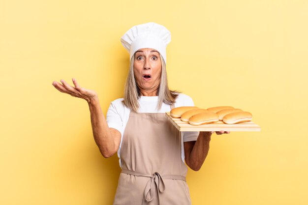 Mulher chef de meia-idade boquiaberta e pasma, chocada e atônita com uma surpresa incrível e segurando uma bandeja de pão