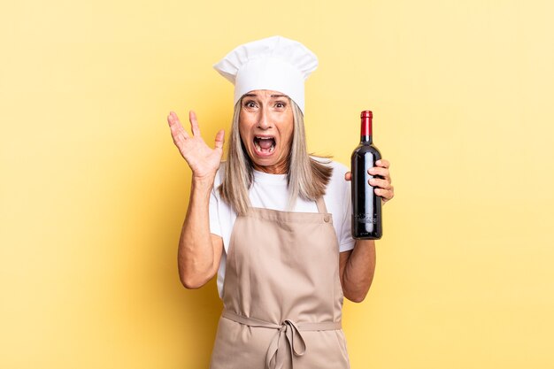 Mulher chef de meia-idade gritando com as mãos para o alto, sentindo-se furiosa, frustrada, estressada e chateada segurando uma garrafa de vinho