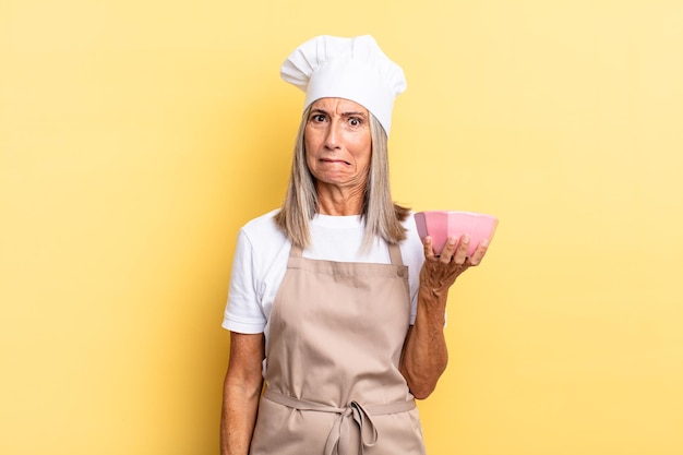 Foto mulher chef de meia-idade parecendo perplexa e confusa, mordendo o lábio com um gesto nervoso, sem saber a resposta para o problema e segurando uma panela vazia