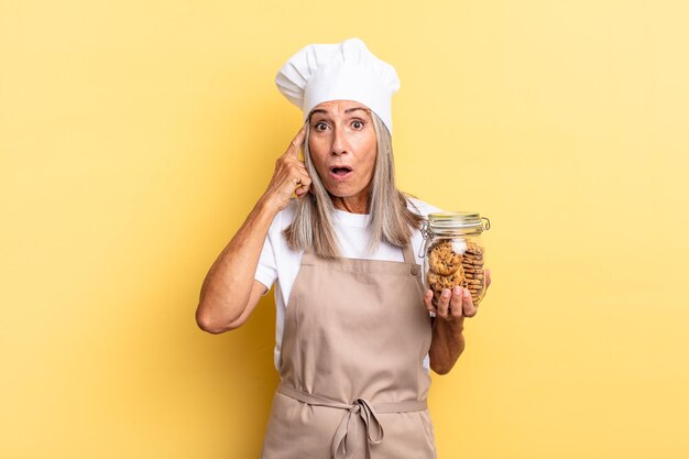 Mulher chef de meia-idade parecendo surpresa, boquiaberta, chocada, percebendo um novo pensamento, ideia ou conceito com biscoitos