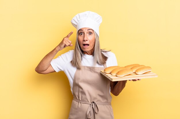 Mulher chef de meia-idade parecendo surpresa, boquiaberta, chocada, percebendo um novo pensamento, ideia ou conceito e segurando uma bandeja de pão