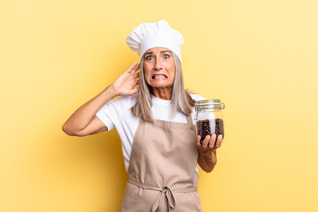 Mulher chef de meia-idade se sentindo estressada, preocupada, ansiosa ou assustada, com as mãos na cabeça, entrando em pânico com o erro segurando grãos de café