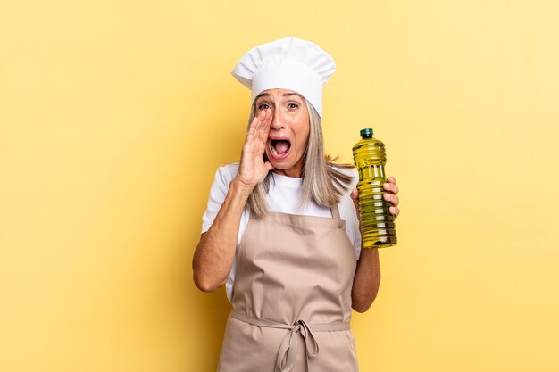 Mulher chef de meia-idade se sentindo feliz, animada e positiva, dando um grande grito com as mãos perto da boca, gritando