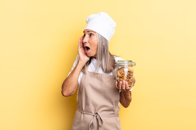 Mulher chef de meia-idade se sentindo feliz, animada e surpresa, olhando para o lado com as duas mãos no rosto com biscoitos