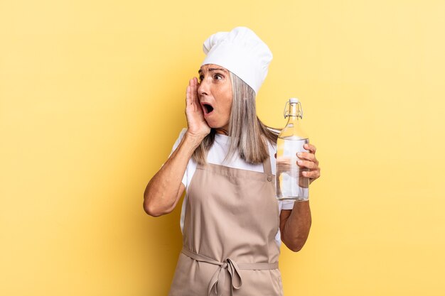 Mulher chef de meia-idade se sentindo feliz, animada e surpresa, olhando para o lado com as duas mãos no rosto com uma garrafa de água