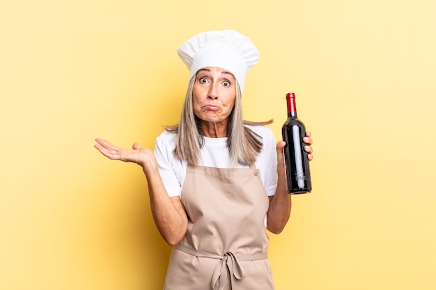 Mulher chef de meia-idade se sentindo perplexa e confusa, duvidando, ponderando ou escolhendo opções diferentes com uma expressão engraçada segurando uma garrafa de vinho