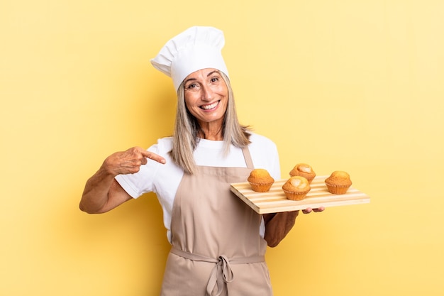 Mulher chef de meia-idade sorrindo alegremente, sentindo-se feliz e apontando para o lado e para cima, mostrando o objeto no espaço da cópia e segurando uma bandeja de muffins