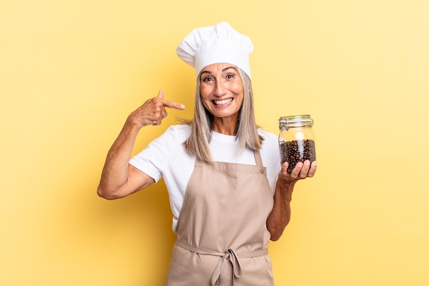 Mulher chef de meia-idade sorrindo com confiança apontando para o próprio sorriso largo, atitude positiva, relaxada e satisfeita segurando grãos de café