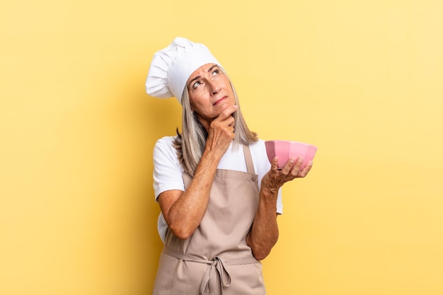 Mulher chef de meia-idade sorrindo com uma expressão feliz e confiante com a mão no queixo, pensando e olhando para o lado e segurando uma panela vazia