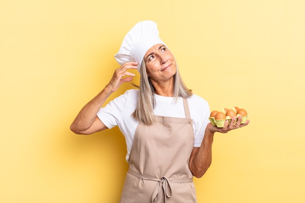 Mulher chef de meia-idade sorrindo feliz e sonhando acordada ou duvidando, olhando para o lado segurando uma caixa de ovos