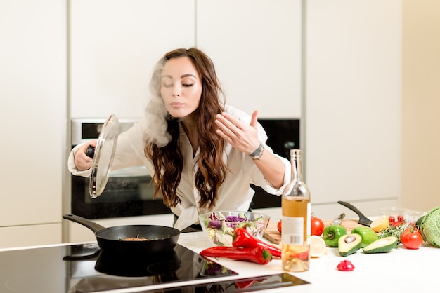 Mulher cheirando vapor da panela e cozinhar peixe fresco com vinho branco e legumes na cozinha