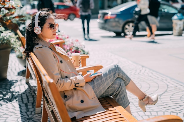 mulher chinesa asiática confiante em óculos de sol, ouvindo música do smartphone com fones de ouvido na rua da cidade. jovem com fone de ouvido desfrutar do sol sentado no banco de madeira segurando o celular.