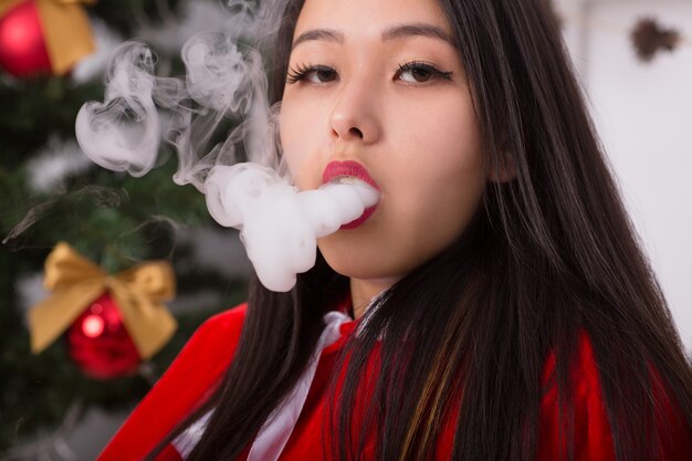 Mulher chinesa com fantasia de Papai Noel fumando um cigarro eletrônico perto do retrato da árvore de Natal