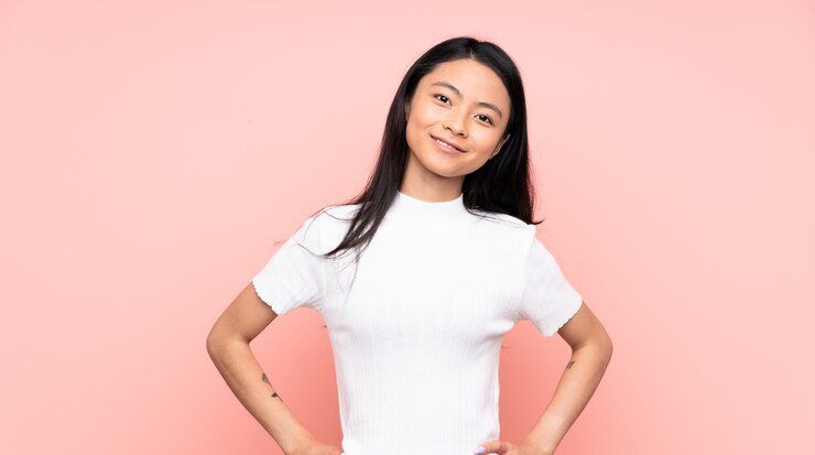 Foto mulher chinesa de adolescente na parede rosa posando com os braços no quadril e sorrindo
