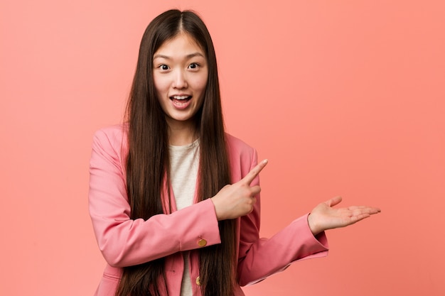 Mulher chinesa de negócios jovem vestindo terno rosa animado segurando um espaço de cópia na palma da mão.