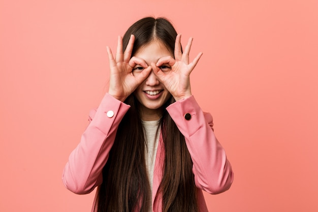 Mulher chinesa de negócios jovem vestindo terno rosa mostrando sinal bem sobre os olhos