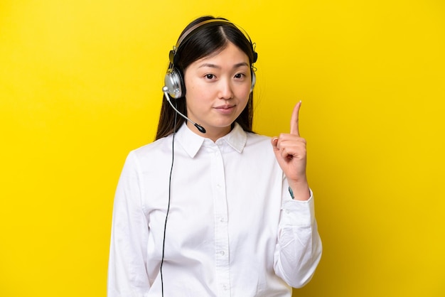 Mulher chinesa de telemarketing trabalhando com um fone de ouvido isolado em fundo amarelo apontando com o dedo indicador uma ótima ideia