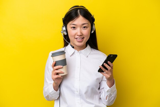 Mulher chinesa de telemarketing trabalhando com um fone de ouvido isolado em fundo amarelo segurando café para levar e um celular