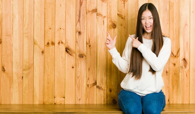 Mulher chinesa nova que senta-se em um lugar de madeira que aponta com o dedo indicador a, expressando o excitamento e o desejo.