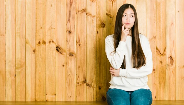 Mulher chinesa nova que senta-se em um lugar de madeira que olha lateralmente com expressão duvidosa e cética.