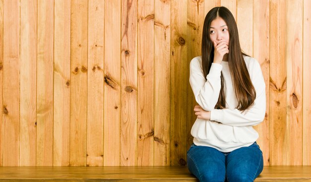 Mulher chinesa nova que senta-se em um lugar de madeira que olha pensativo a um espaço da cópia que cobre a boca com a mão.