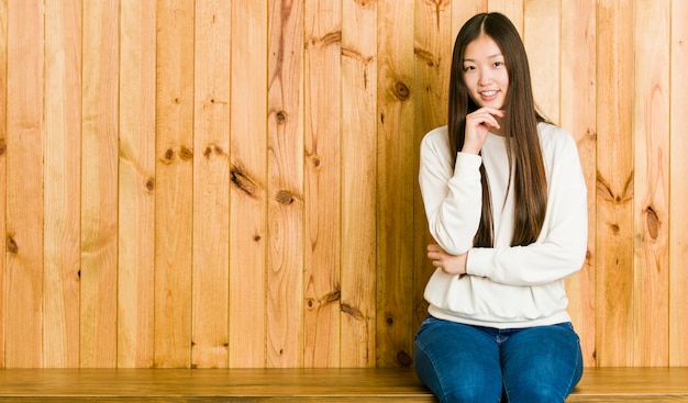 Mulher chinesa nova que senta-se em um lugar de madeira que sorri queixo feliz e seguro, tocante com mão.