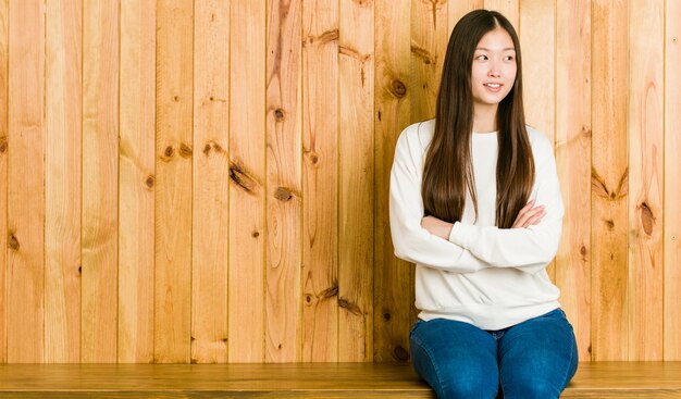 Mulher chinesa nova que senta-se em um lugar de madeira que sorri seguro com braços cruzados.