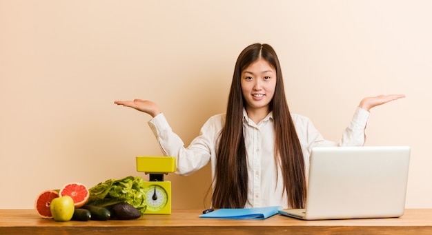 Mulher chinesa nutricionista jovem trabalhando com seu laptop faz escala com os braços, sente-se feliz e confiante.