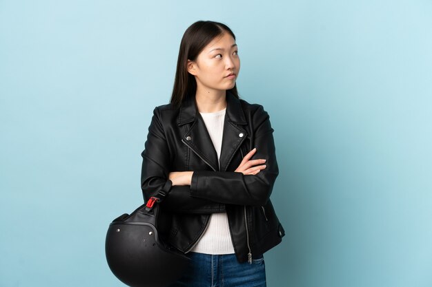 Mulher chinesa segurando um capacete de motocicleta sobre um azul isolado, pensando uma ideia