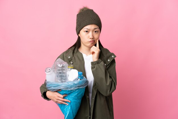 Mulher chinesa, segurando um saco cheio de garrafas de plástico para reciclar sobre parede rosa isolada e pensando