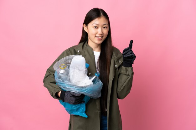 Mulher chinesa, segurando um saco cheio de garrafas de plástico para reciclar sobre parede rosa isolada, mostrando e levantando um dedo em sinal dos melhores