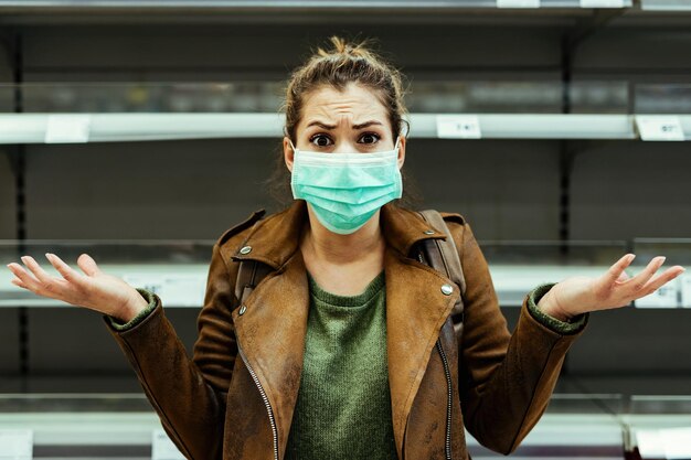 Foto mulher chocada com máscara protetora gesticulando em frente a prateleira vazia no supermercado durante pandemia de coronavírus