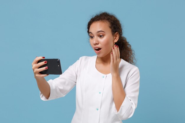 Mulher chocada do médico afro-americano isolada no fundo azul. Médico em vestido médico fazendo selfie filmado no celular faz videochamada. Conceito de medicina pessoal de saúde. Simule o espaço da cópia.