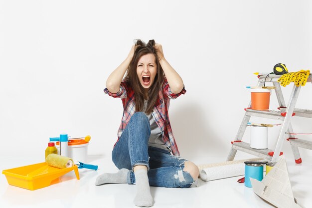 Mulher chocada louca gritando confusa, sentada no chão com instrumentos para apartamento de renovação isolado no fundo branco. Papel de parede, acessórios para colar, ferramentas de pintura. Conceito de reparo em casa.