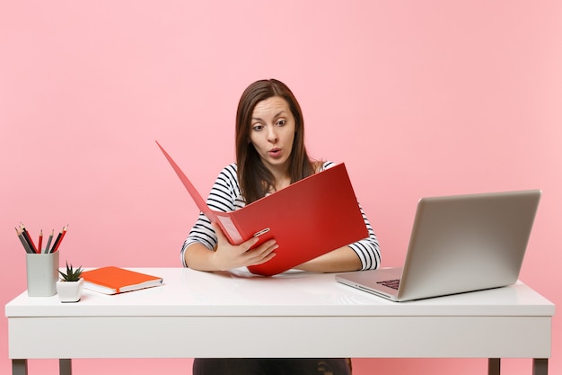 Mulher chocada olhando na pasta vermelha com documentos em papel e trabalhando no projeto enquanto está sentado no escritório com o laptop isolado no fundo rosa pastel. Conceito de carreira empresarial de realização. Copie o espaço.