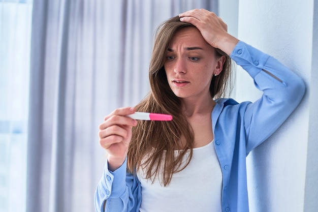 Mulher chocada segurando a cabeça olha para o resultado positivo do teste de gravidez