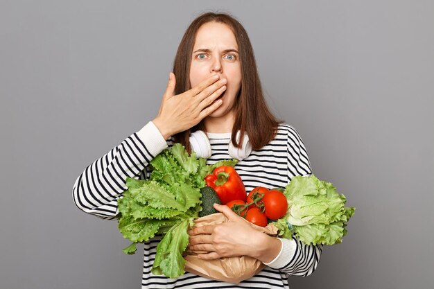 Mulher chocada segurando legumes cobrindo a boca com a mão isolada sobre fundo cinza Comer fresco ficar saudável Benefícios de bem-estar de alimentos orgânicos