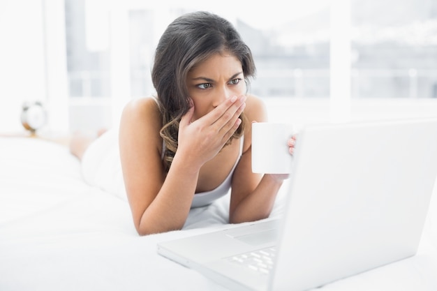 Mulher chocada tomando café ao usar o laptop na cama