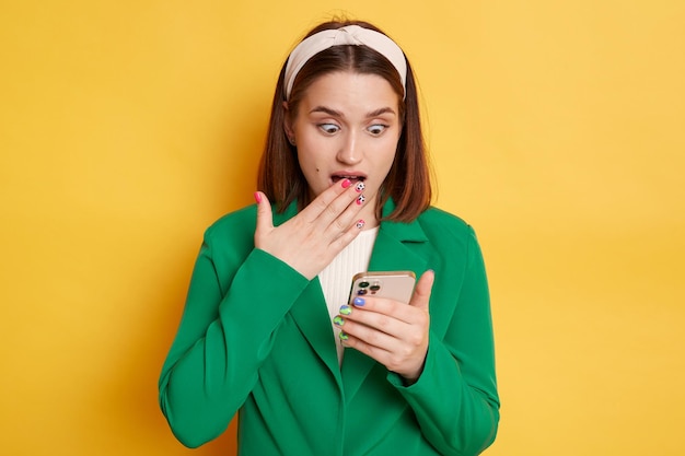 Mulher chocada vestindo jaqueta verde posando isolada sobre fundo amarelo usando telefone celular lendo notícias nas redes sociais