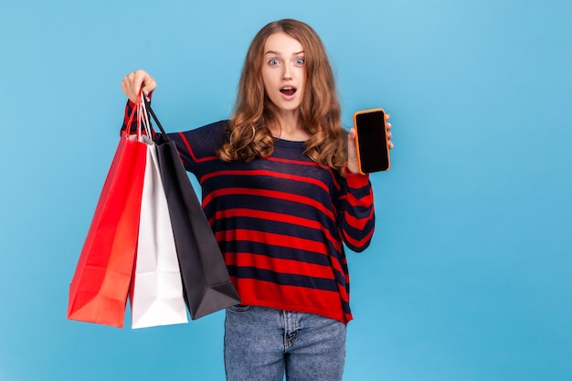 Mulher chocada vestindo suéter listrado estilo casual em pé segurando o celular com display vazio para promoção e sacolas de compras Estúdio interior isolado em fundo azul